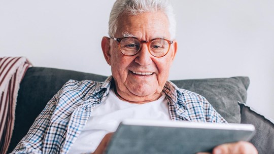 A close up of an old man in his 70s sitting on the couch at home, searching the Internet on his digital tablet, enjoying