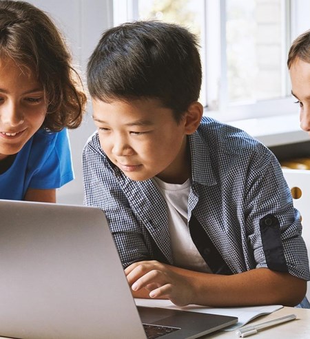 Diverse happy school kids using laptop computer together in classroom. Multicultural children junior students classmates learning online elementary education program class gathered at desk.